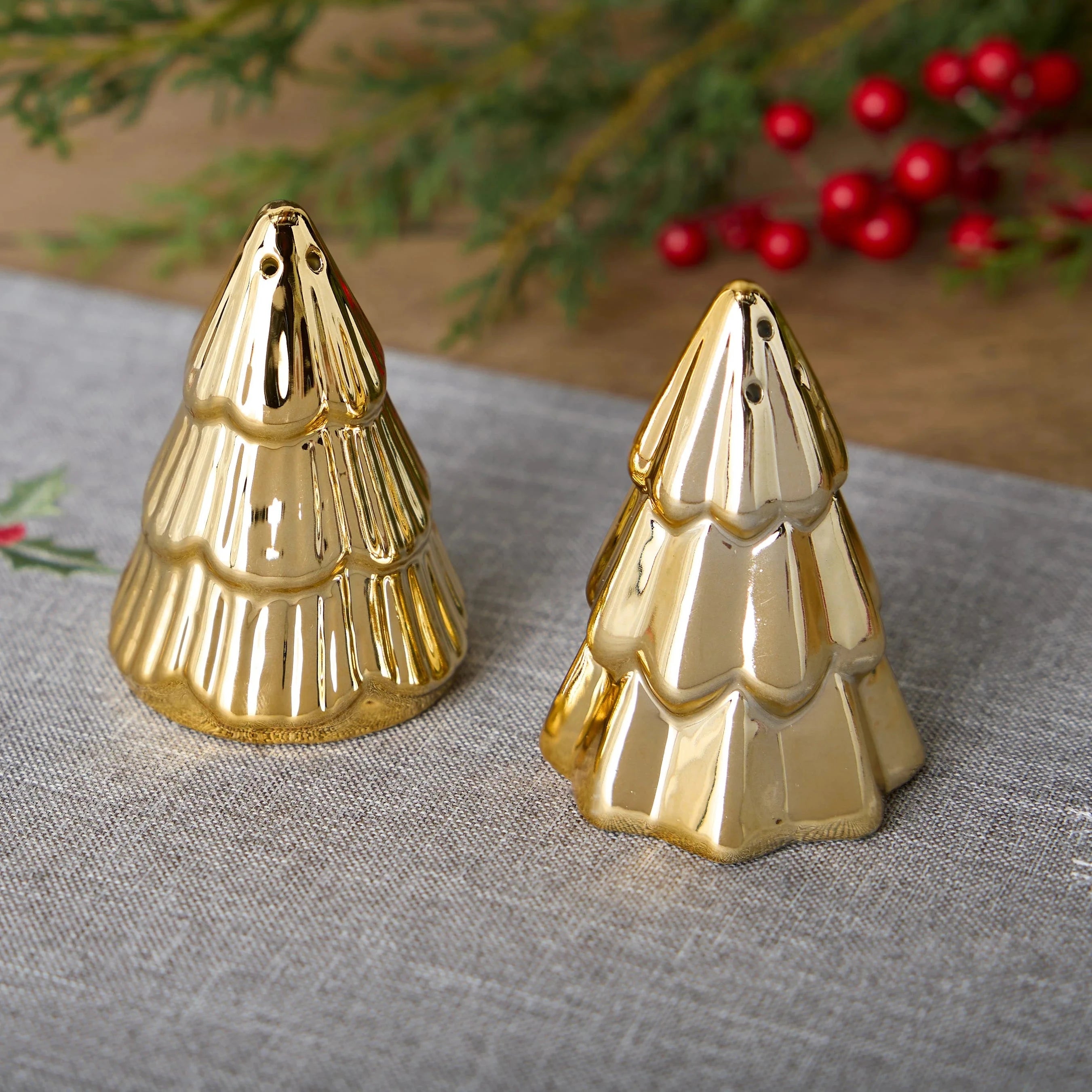 Two gold Christmas tree-shaped ornaments on a textured surface with blurred greenery and red berries in the background.
