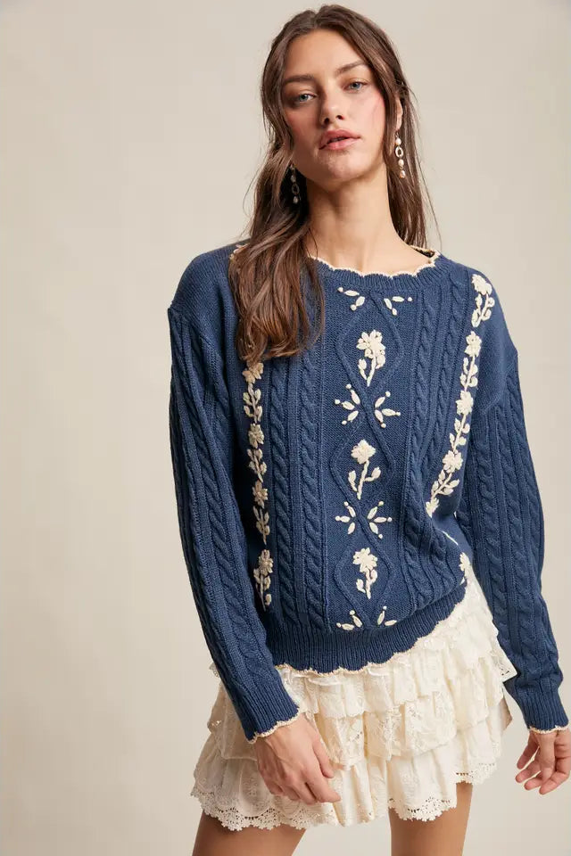 Woman wearing a blue cable knit sweater with floral embroidery on a beige background