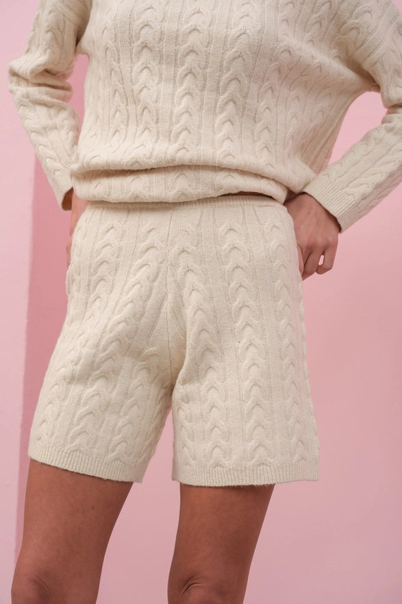 Person wearing a cream cable knit sweater and shorts on a pink background