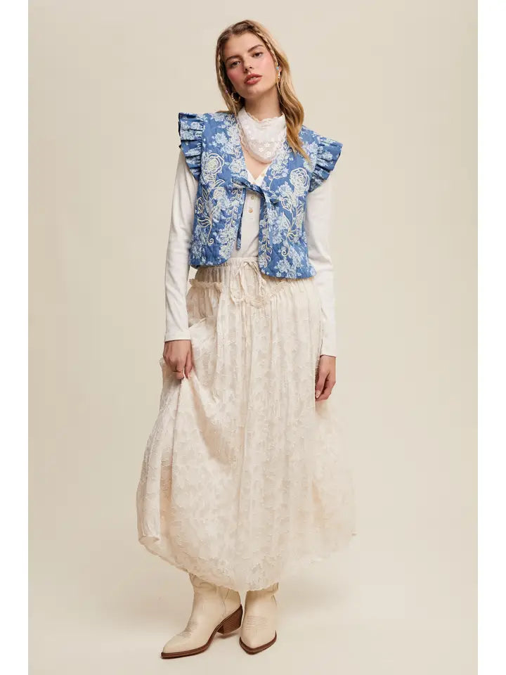 Woman wearing a blue floral vest over a white blouse and beige skirt on a plain background