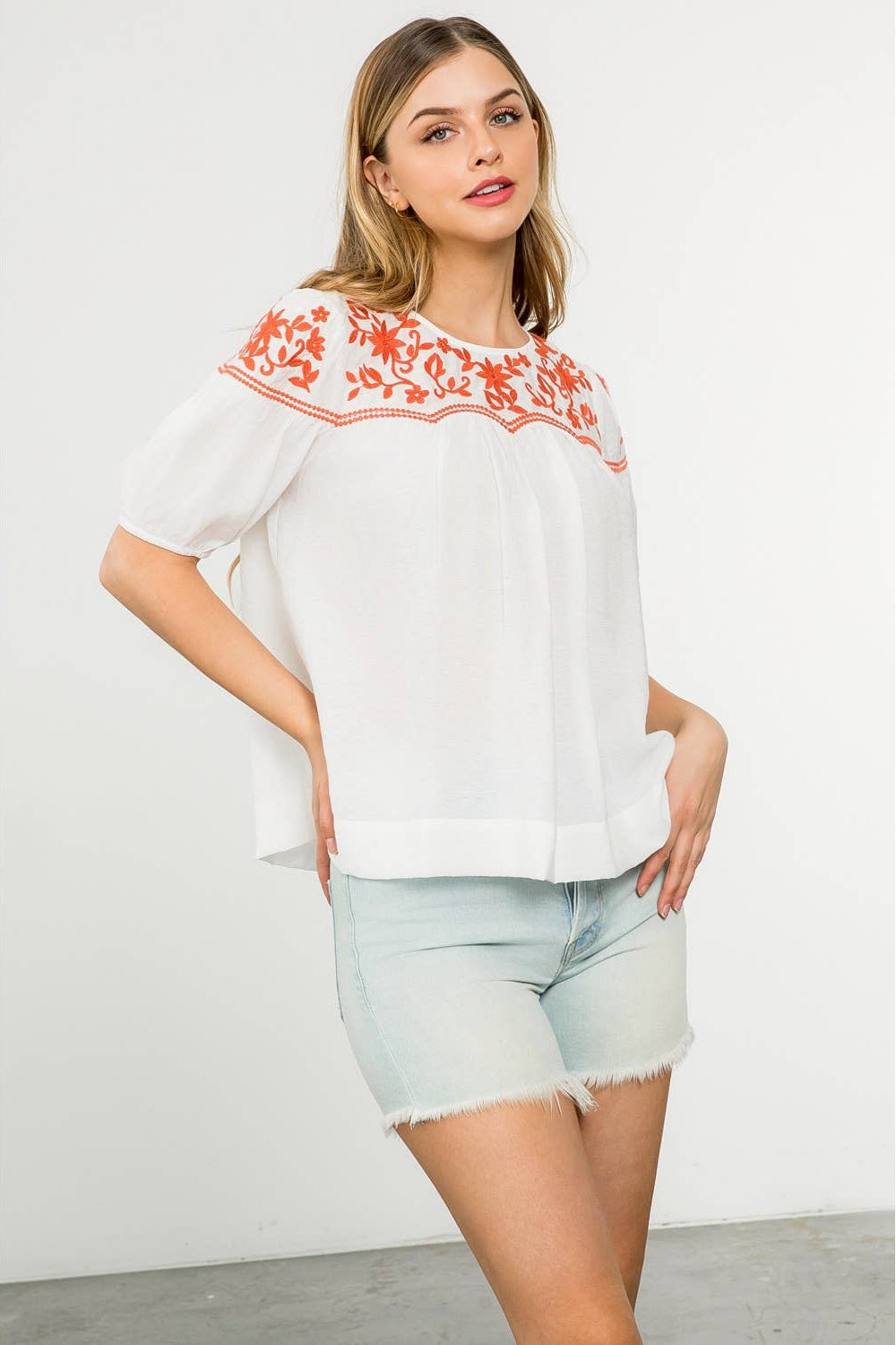 Woman wearing a white blouse with orange embroidery and light blue shorts on a white background