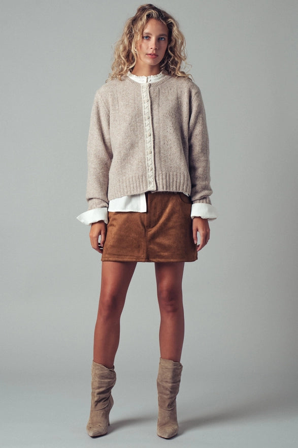 Woman wearing a beige sweater, white shirt, brown skirt, and brown boots on a gray background