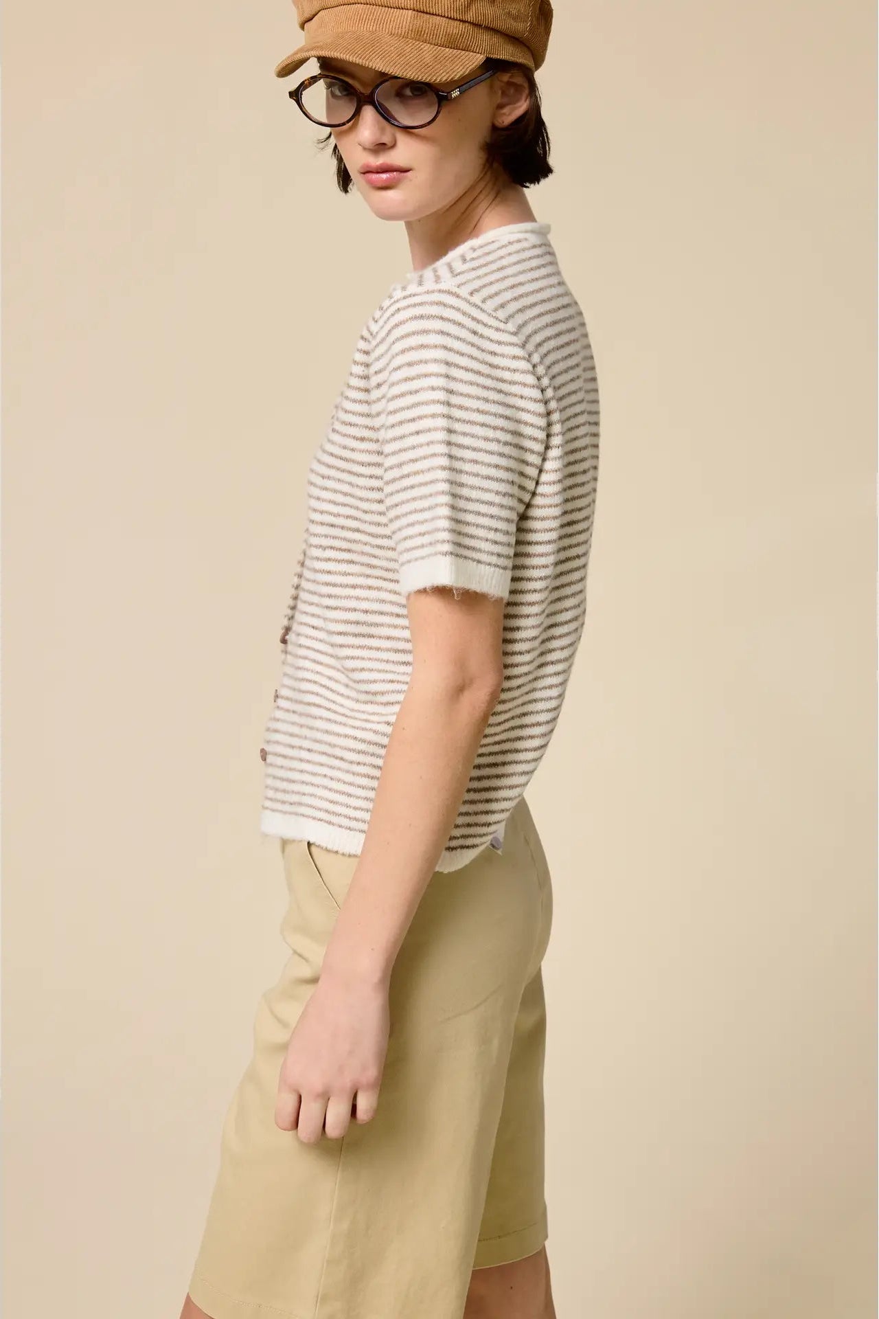 Person wearing a striped shirt, beige cap, and glasses against a plain background