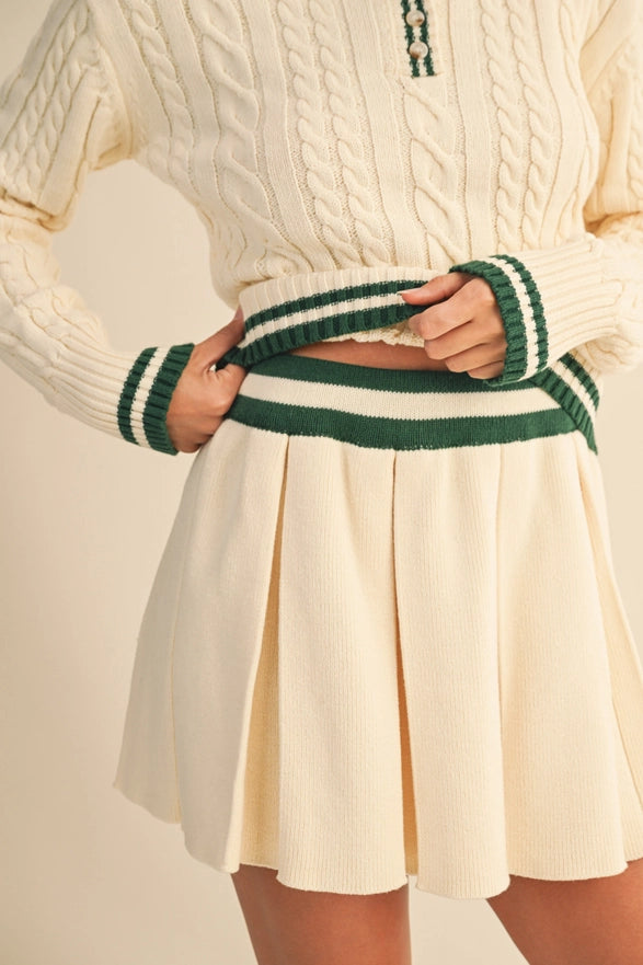 Person wearing a cream sweater with green trim and a pleated cream skirt on a plain background