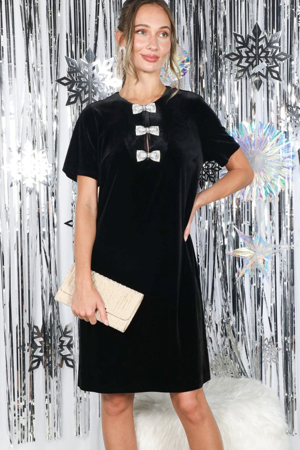 Woman wearing a black dress with bow details against a decorative silver and white background.