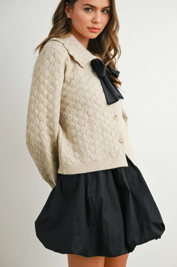 Woman wearing a cream sweater with a black bow and black skirt on a plain background