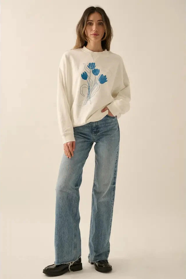 Woman wearing a white sweater with blue floral design and blue jeans on a beige background