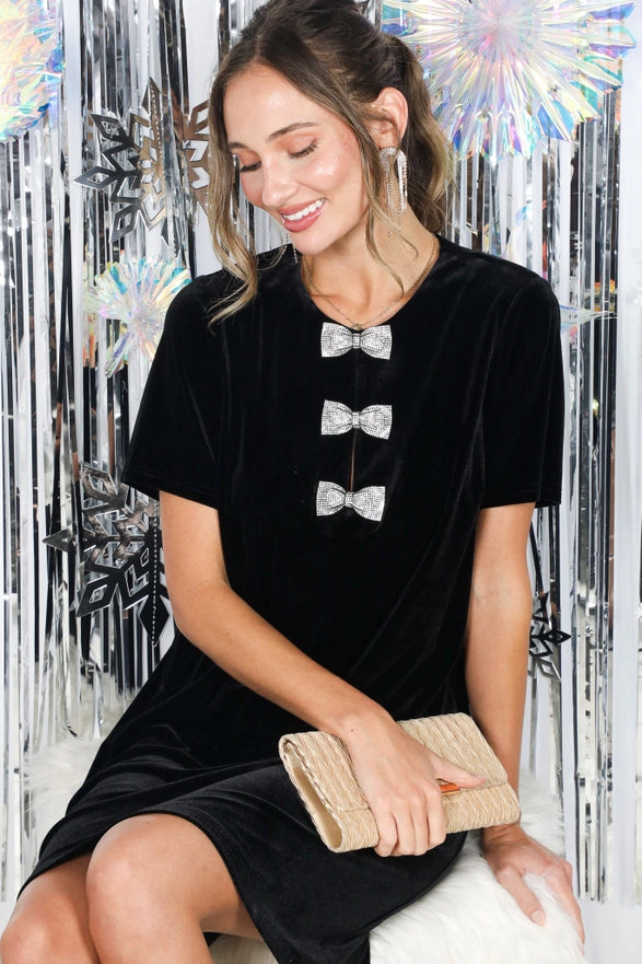 Woman in a black dress with bow details holding a clutch against a decorative silver and white background