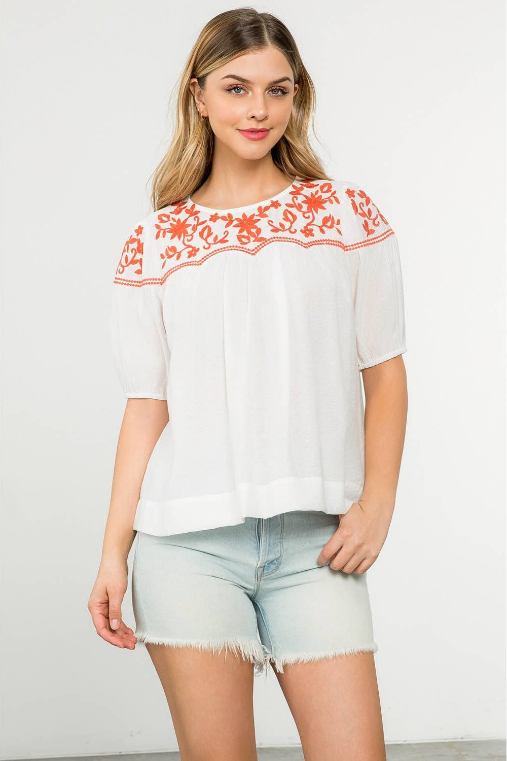 Woman wearing a white blouse with red floral embroidery and light blue denim shorts on a white background