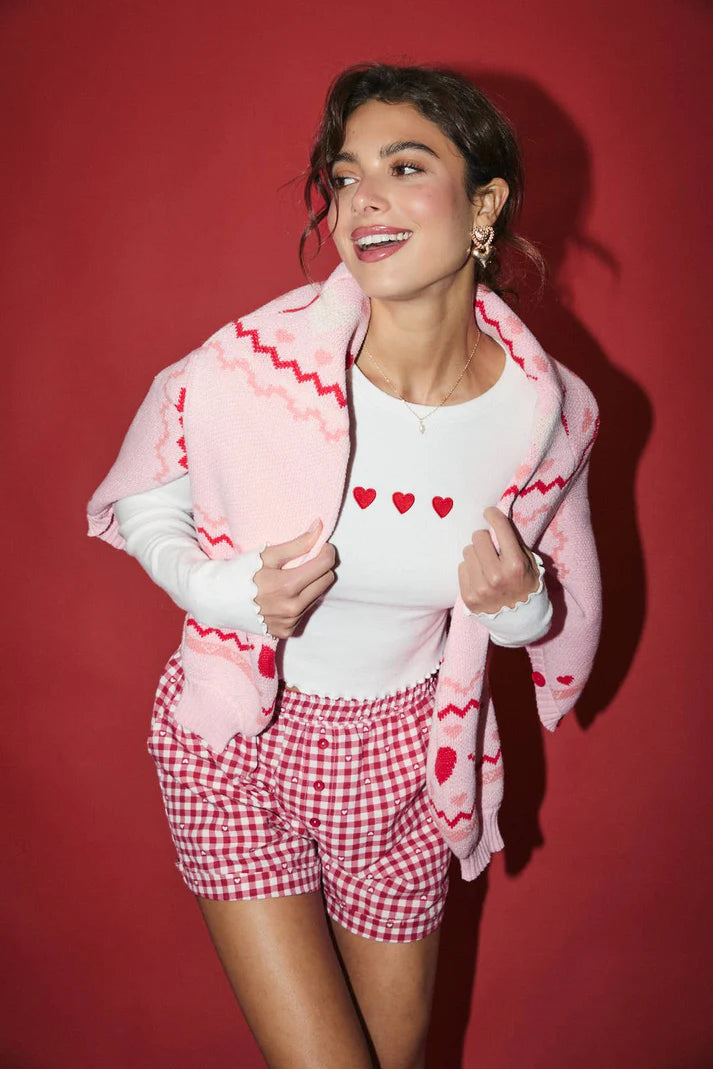 Woman wearing a white shirt with red heart patterns, red checkered shorts, and a pink patterned cardigan against a red background.