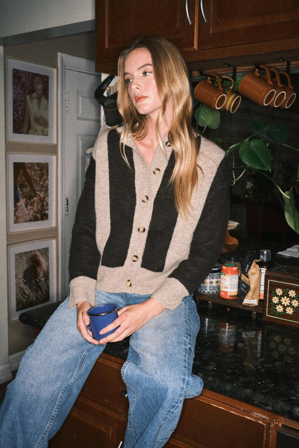 Woman sitting on a kitchen counter holding a blue mug, wearing a striped cardigan and jeans.