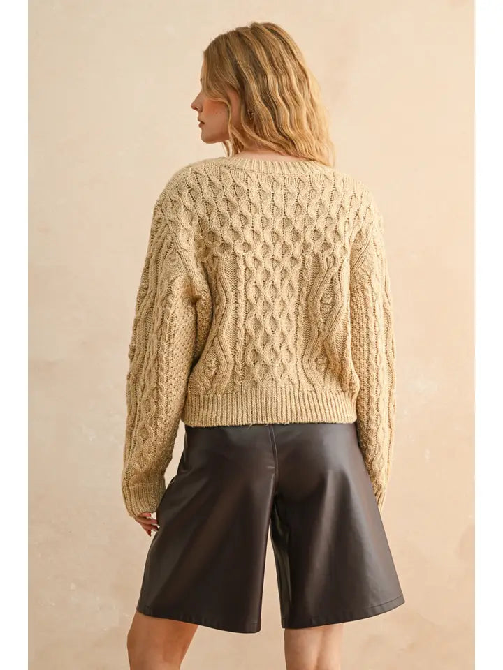 Woman wearing a beige cable knit sweater and black shorts against a beige background