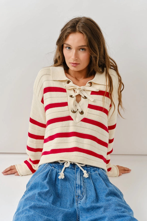 Woman wearing a red and white striped sweater with a lace-up front, sitting against a plain background.