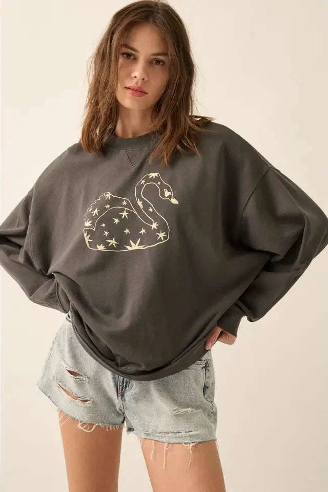 A female model wearing a gray sweatshirt with a vintage-style swan covered in stars graphic print, paired with denim shorts.