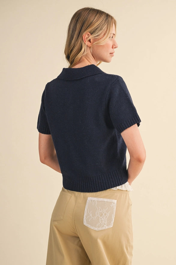 Woman wearing a navy blue short-sleeve sweater over a white shirt with beige pants on a beige background