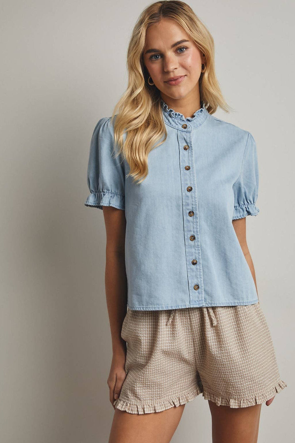 Woman wearing a light blue denim shirt and beige shorts against a plain background