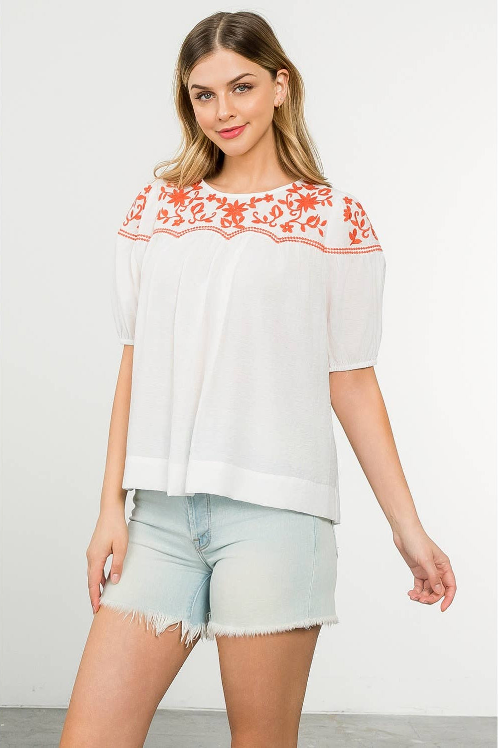 Woman wearing a white blouse with red floral embroidery and light blue shorts on a white background