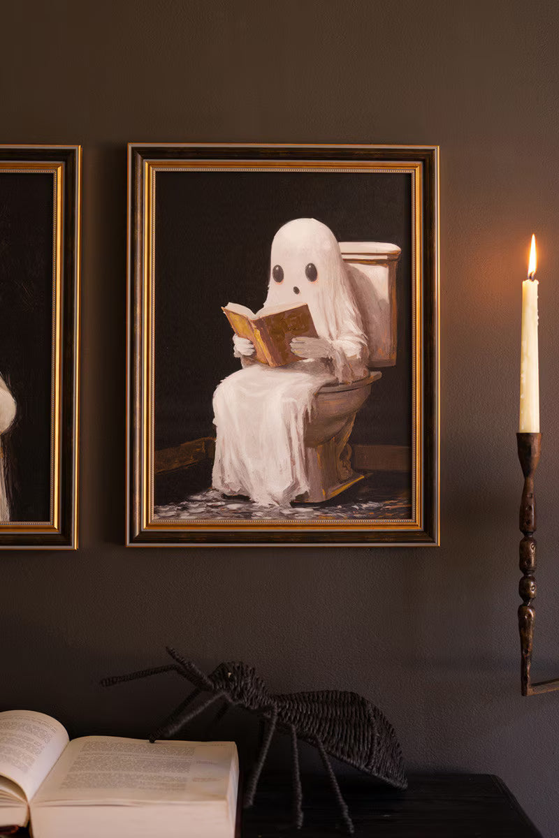 Framed artwork of a ghost reading a book on a toilet against a dark wall with a candle.