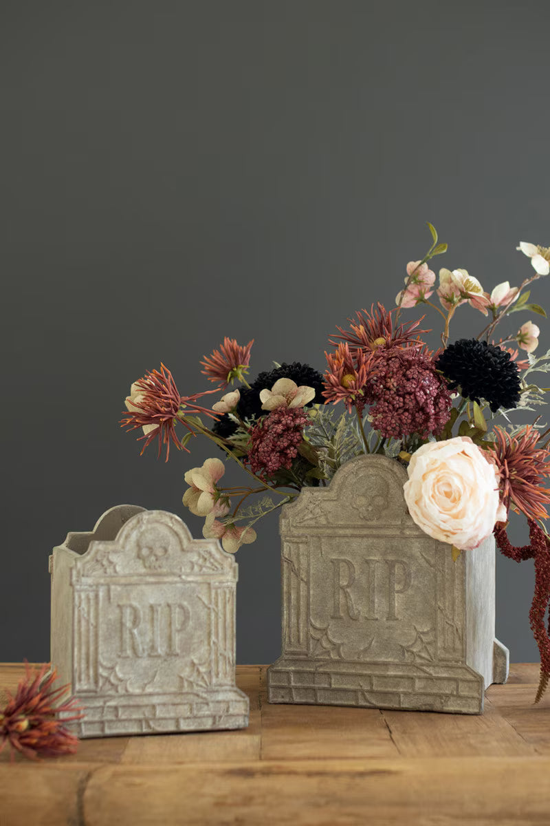Two gray tombstone-shaped decorative vases on a wooden surface, one filled with dried and fresh flowers against a dark gray background.