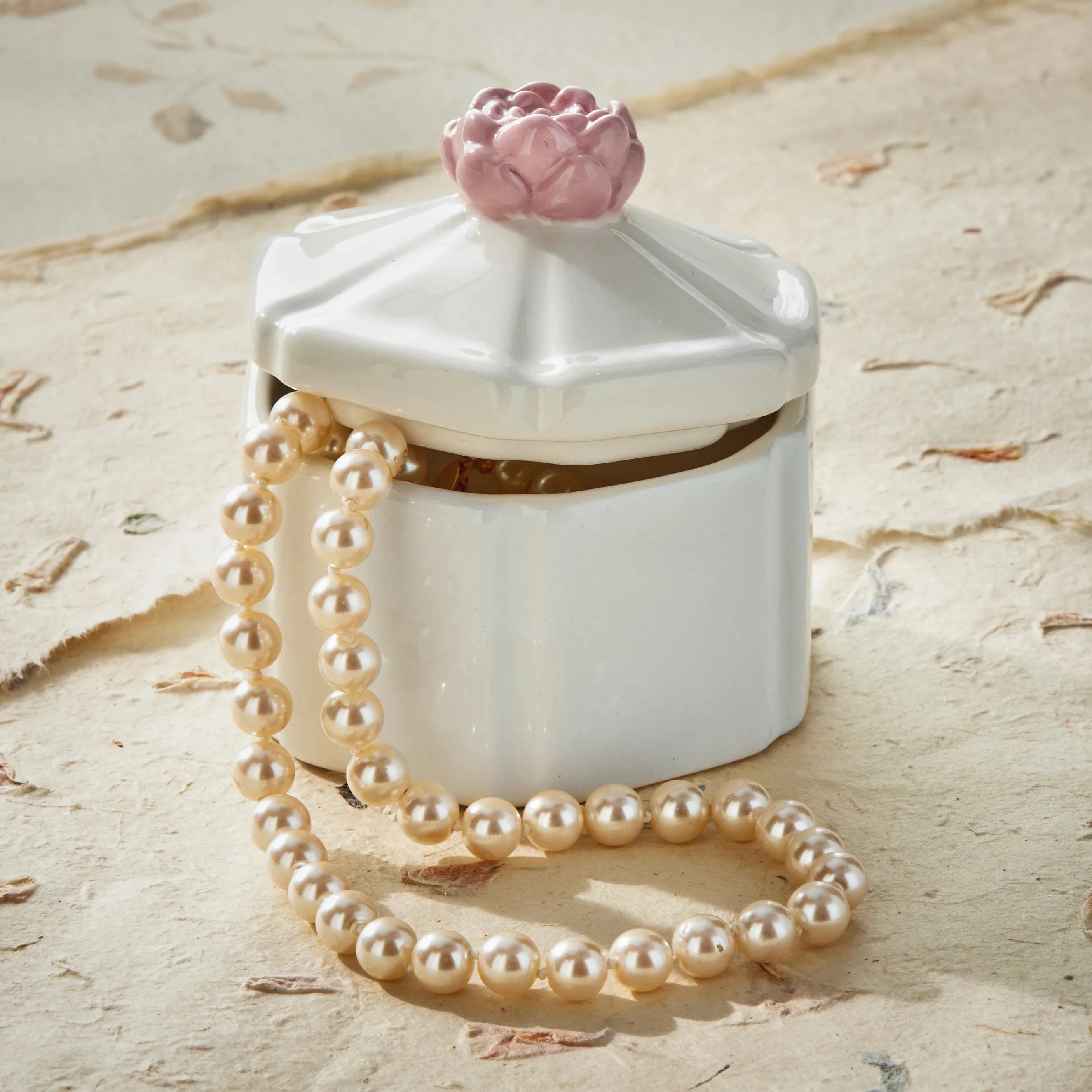 White hexagonal box with pink flower on top and pearl necklace on a textured beige surface