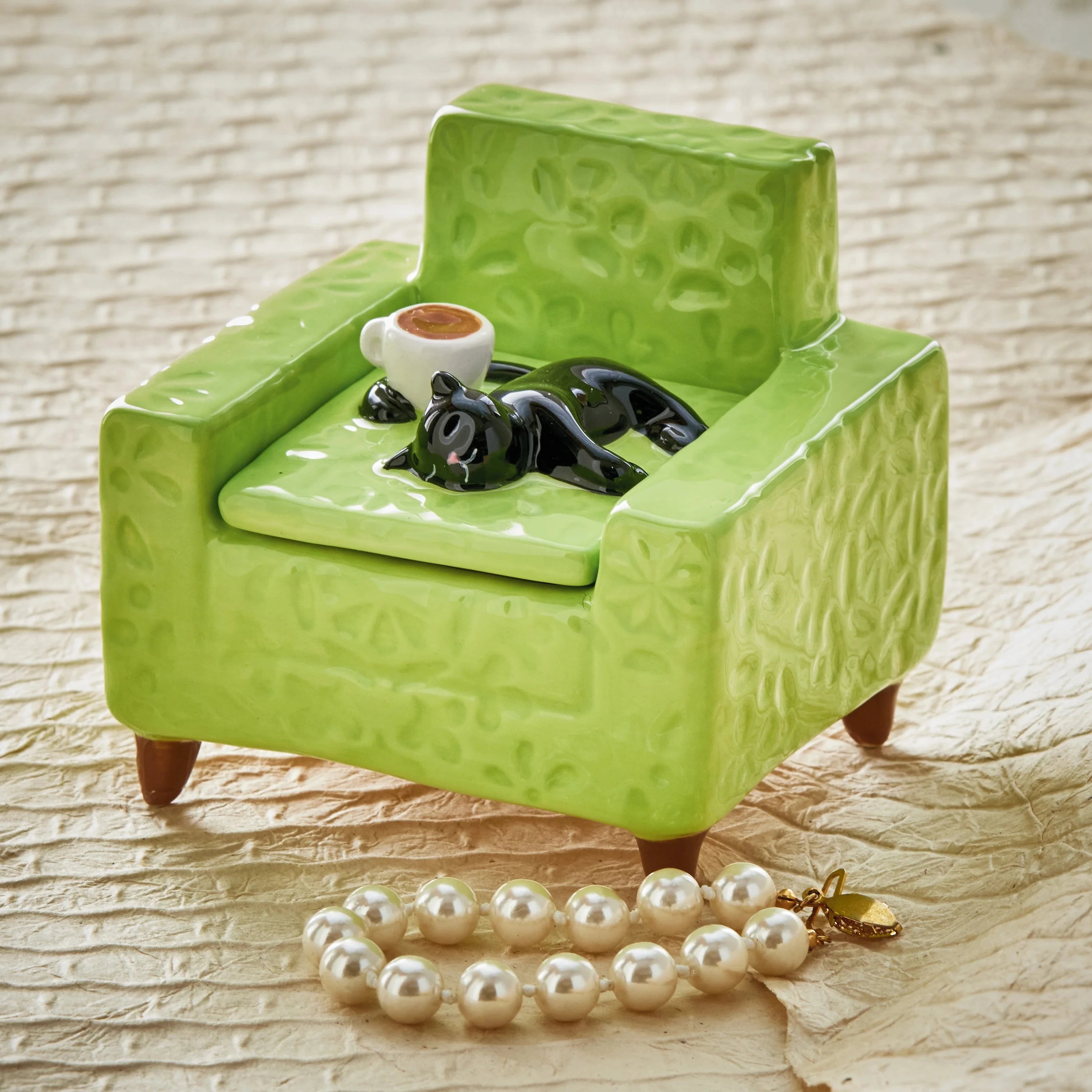 Green textured armchair with a black cat figurine and coffee cup, pearl necklace on beige fabric background