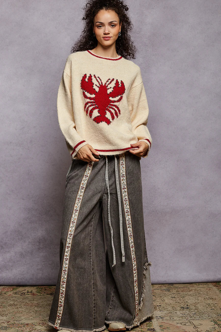 Woman wearing a cream sweater with a red lobster design and gray pants against a gray background