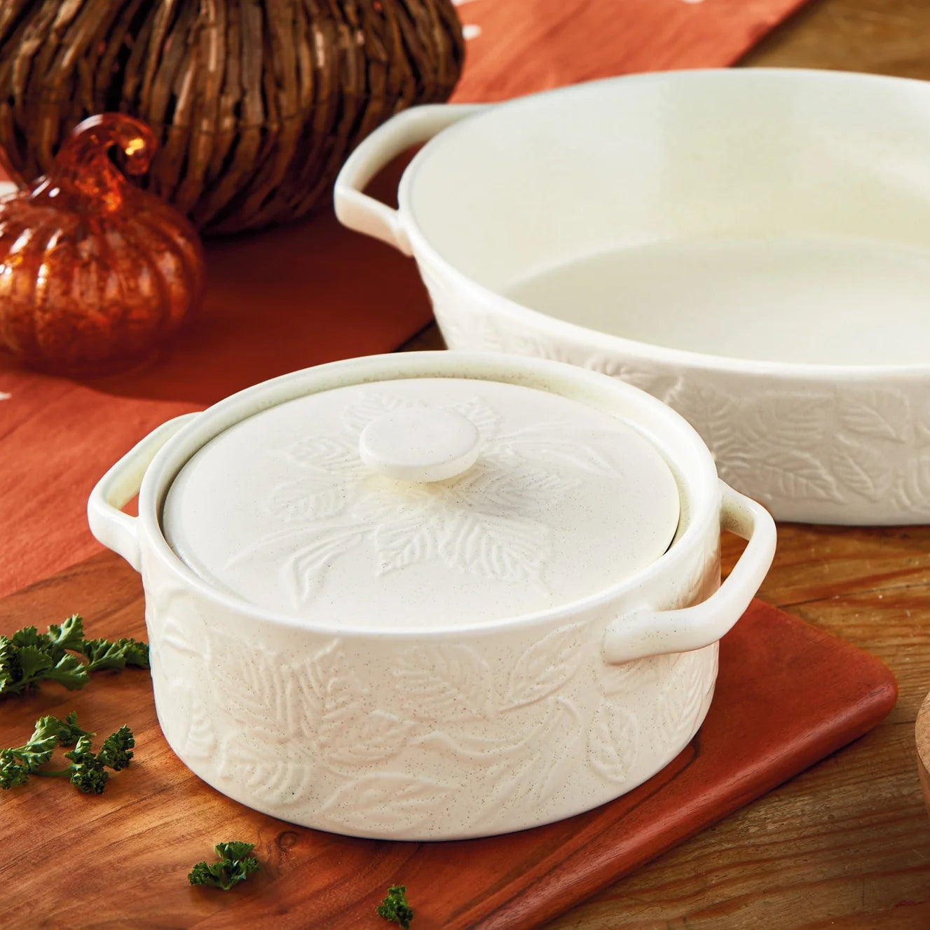 Two white embossed ceramic cookware pieces on a wooden table: a small lidded casserole dish and a larger oval baking dish, styled with peppers, herbs, and autumn decor.