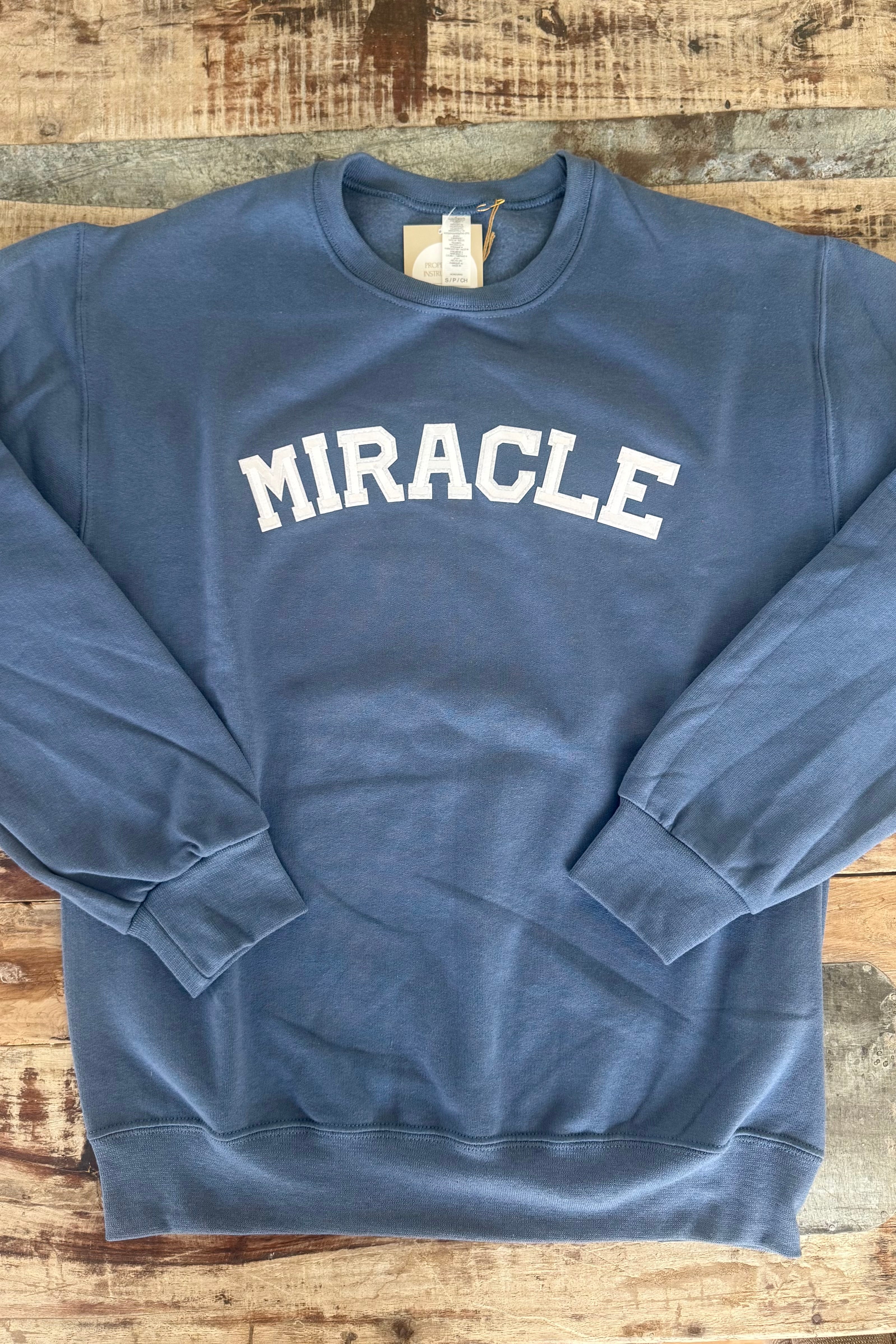Blue sweatshirt with 'MIRACLE' text on a wooden surface, Travis Kelce sweatshirt