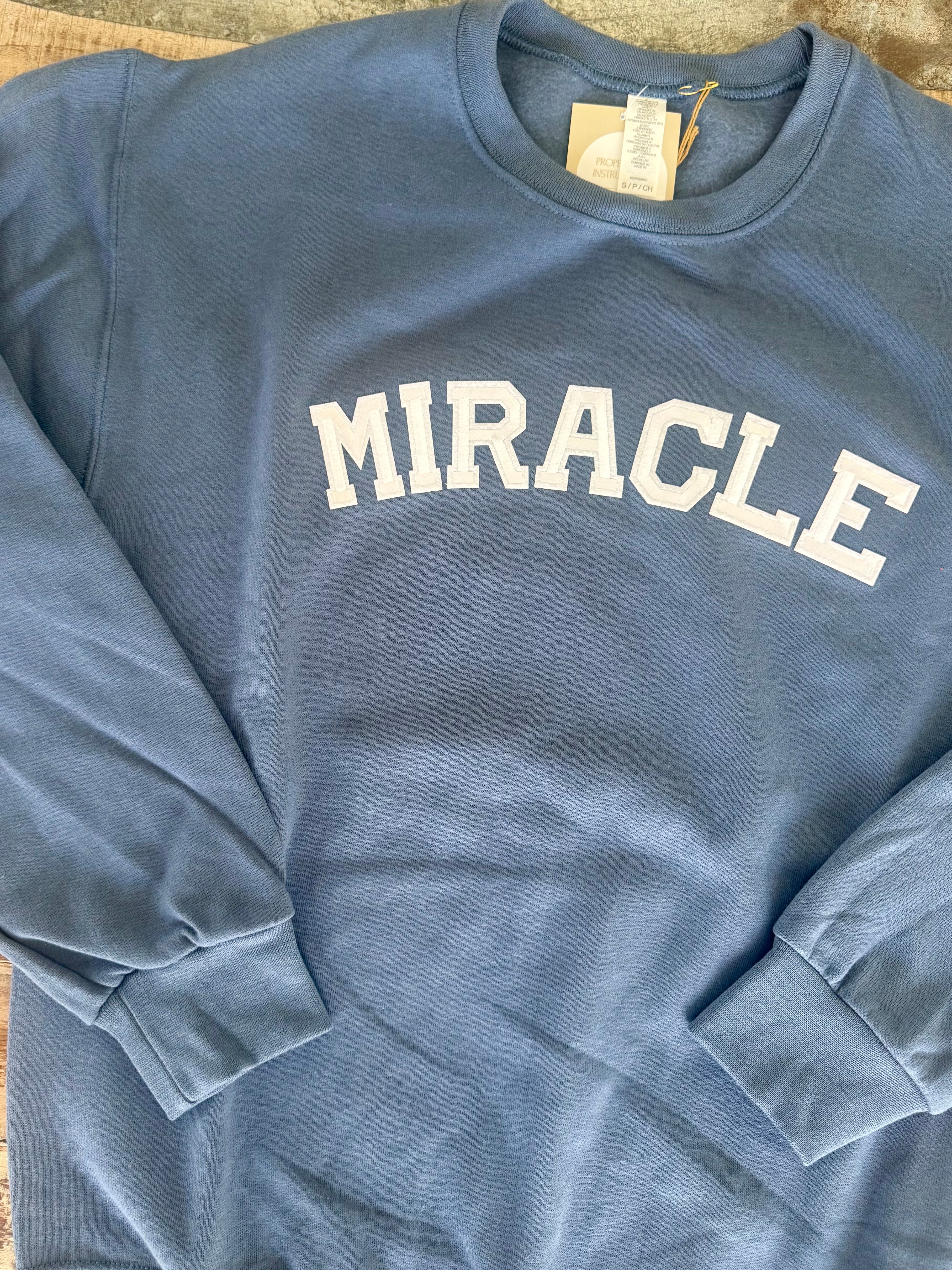 Blue sweatshirt with 'MIRACLE' text on a wooden surface, Travis Kelce sweatshirt