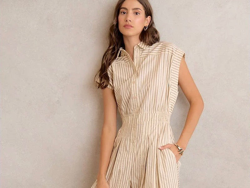 A woman wearing a sleeveless beige and cream vertically striped button-up shirt dress, standing against a textured light gray wall.