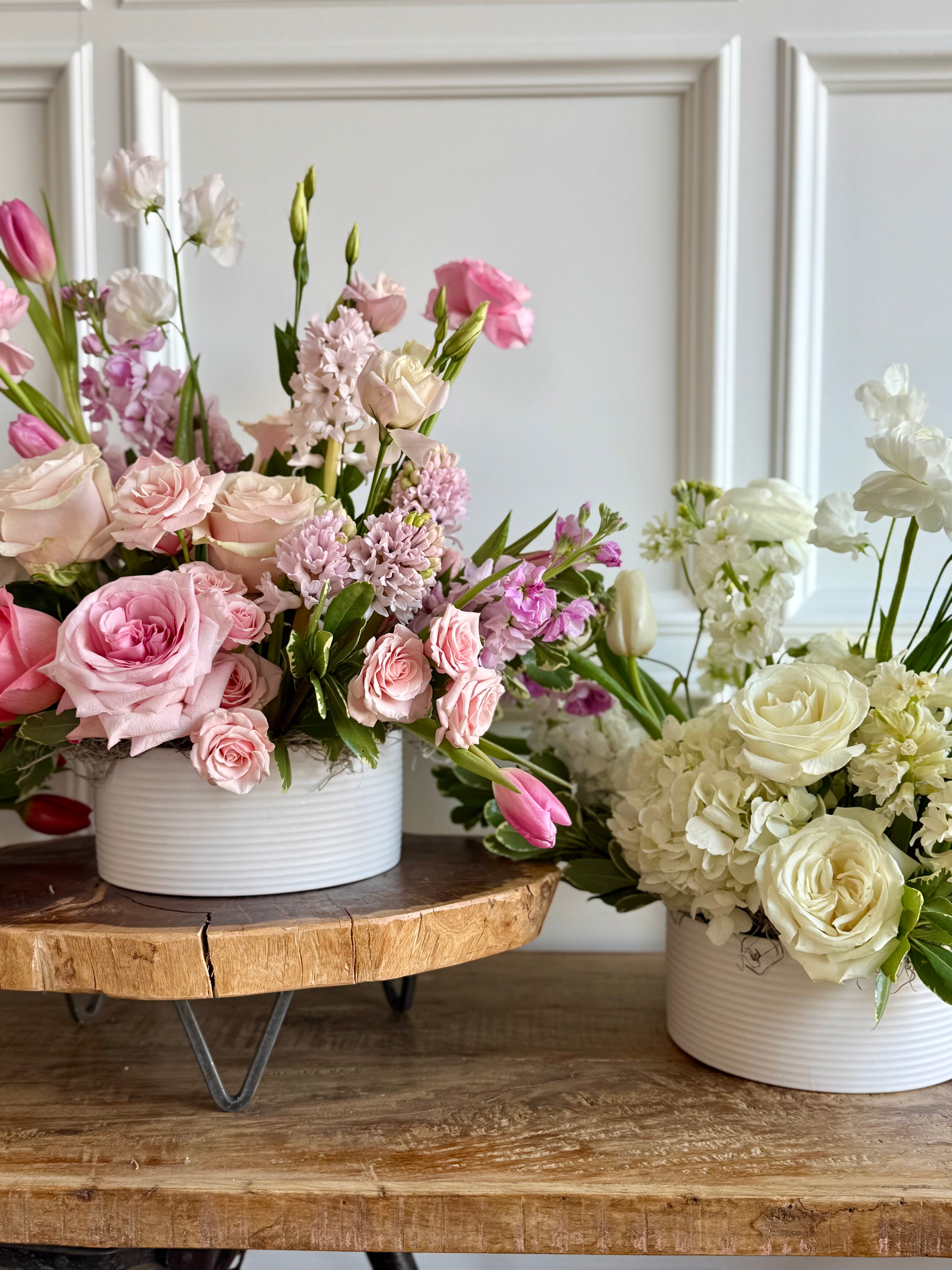 Two flower arrangements in white pots on a wooden table with a neutral background