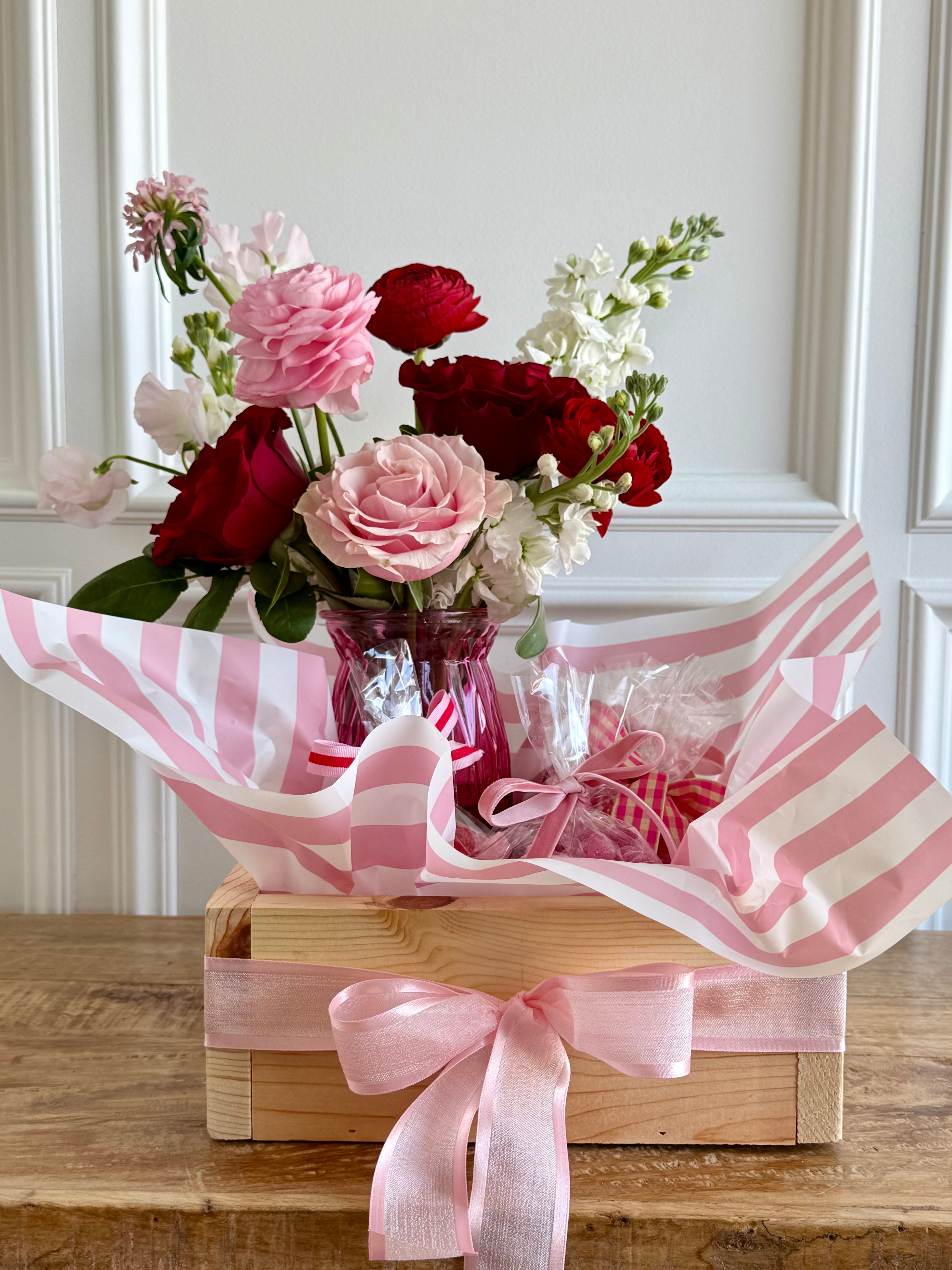 Bouquet of flowers in a wooden box with pink and white striped paper and ribbon on a wooden surface.