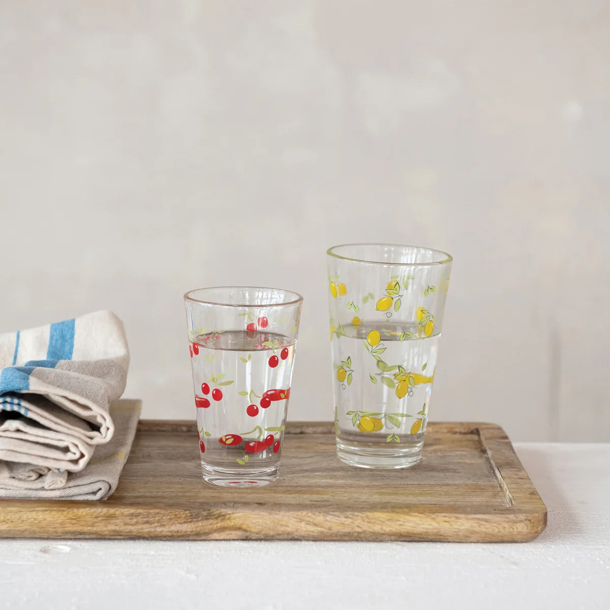 Two glasses with fruit patterns on a wooden tray with a white background