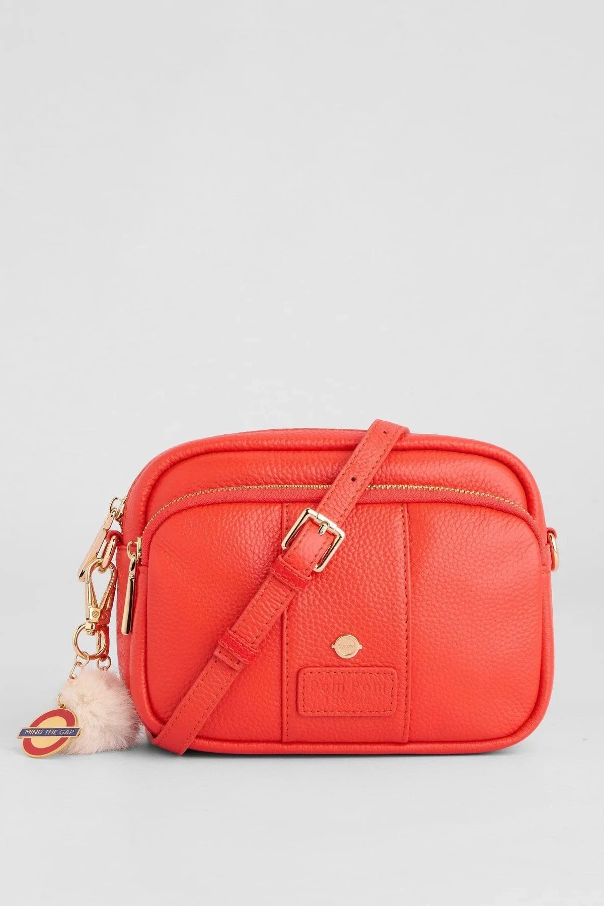 A bright coral-red pebbled leather crossbody bag with a matching adjustable shoulder strap, gold-tone hardware, and a small fluffy cream keychain charm, shown on a light gray background.