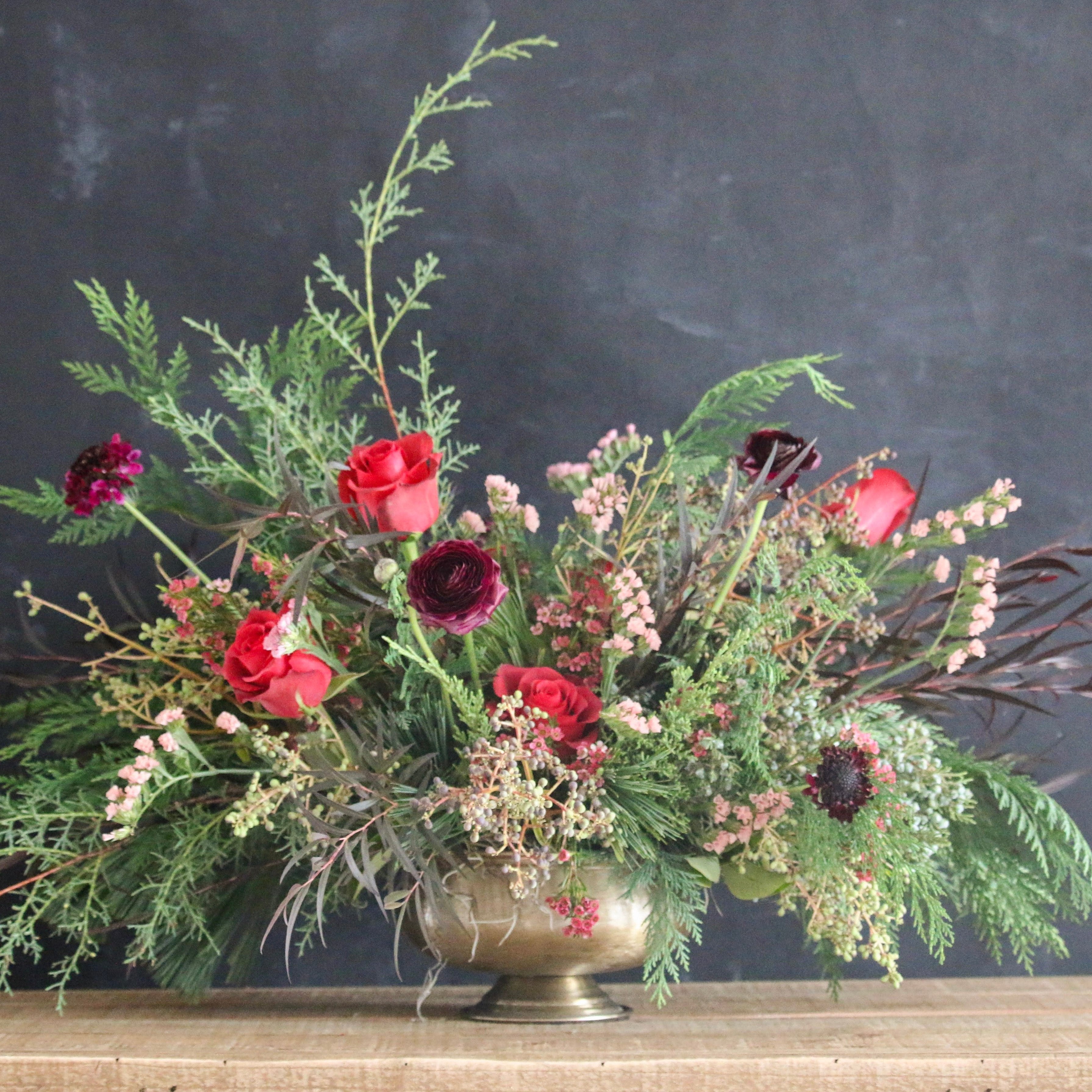 A floral arrangement featuring red, pink, and white flowers with green foliage in a bronze-colored vase, placed on a wooden surface with a dark background.