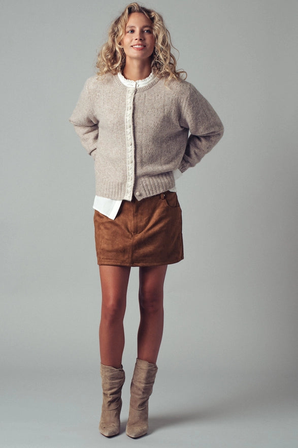 Woman wearing a beige cardigan, white shirt, brown suede skirt, and brown boots on a gray background