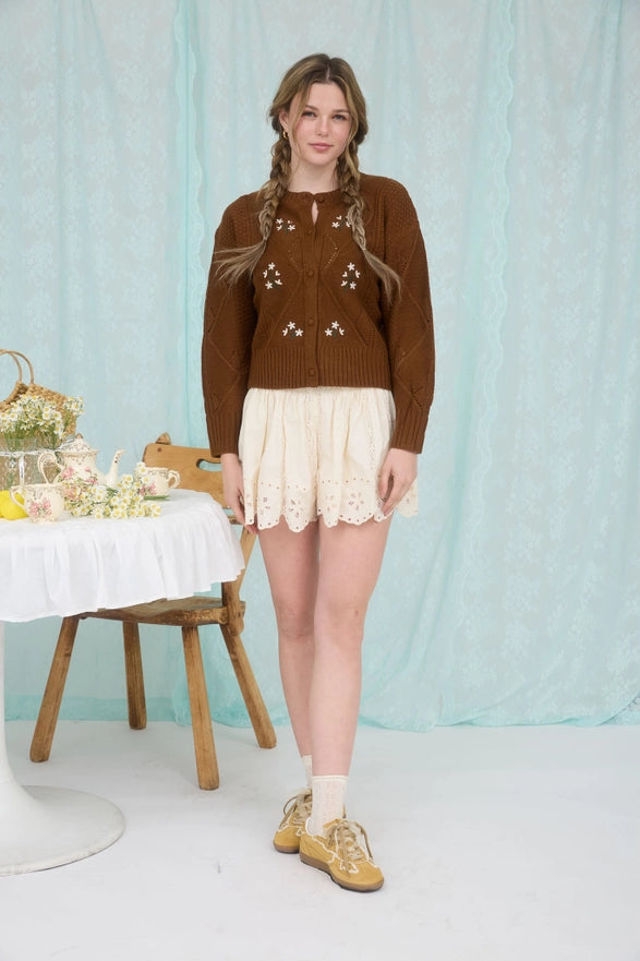 Woman wearing a brown knitted cardigan and white skirt in a studio setting with a light blue curtain.