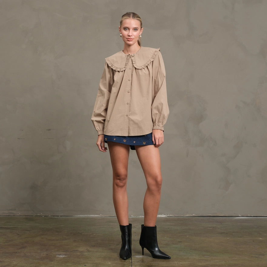 Woman wearing a beige blouse with ruffled details, blue shorts, and black boots against a gray background