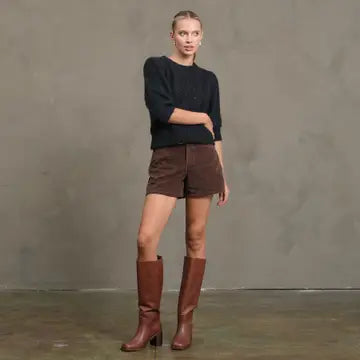 Woman wearing a black sweater and brown shorts with knee-high brown boots against a gray background