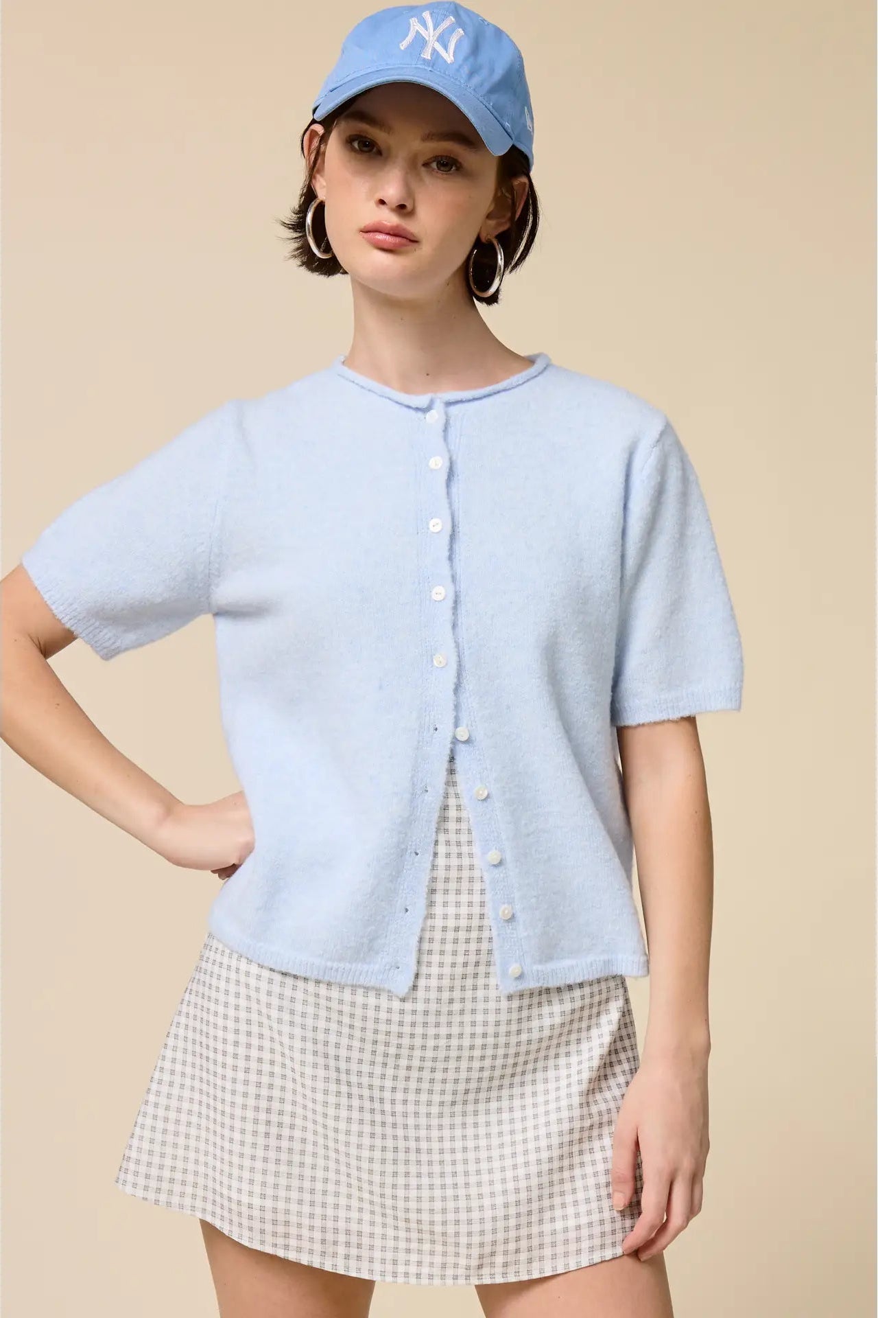 Woman wearing a light blue button-up shirt and checkered skirt with a blue cap on a beige background