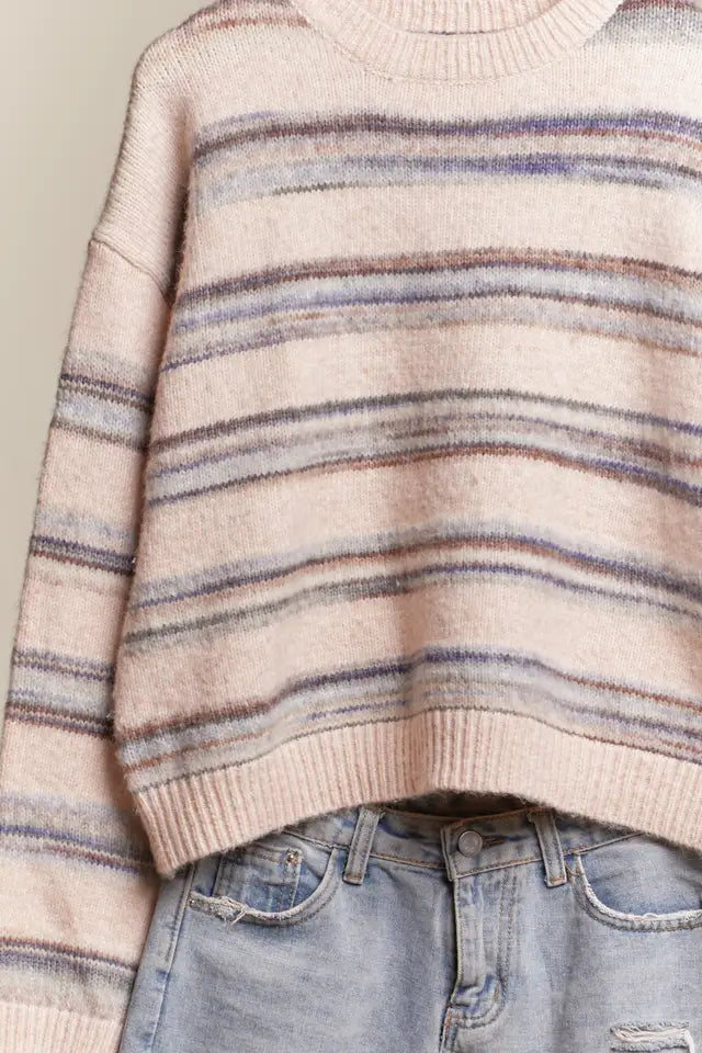 Beige sweater with multicolor stripes worn over blue jeans on a neutral background