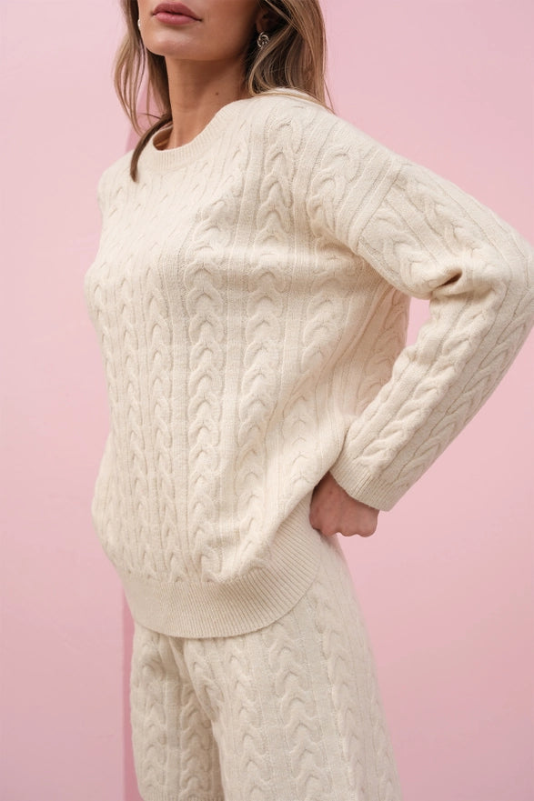 Person wearing a cream cable knit sweater and pants against a pink background