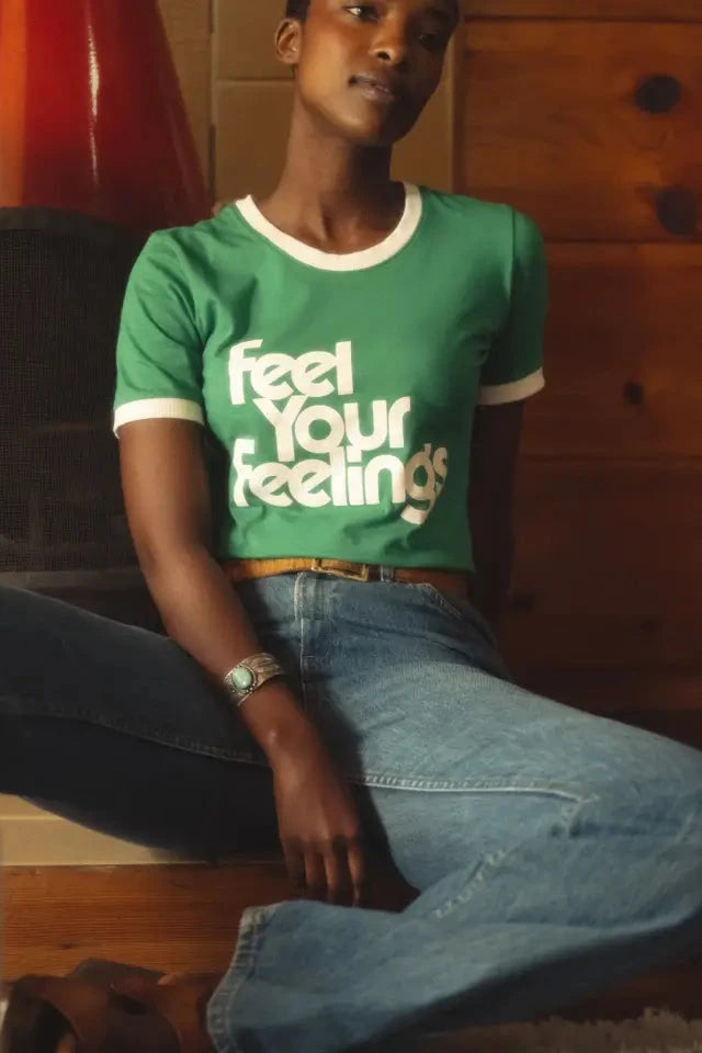 Person wearing a green t-shirt with 'Feel Your Feelings' text, sitting on a wooden floor.