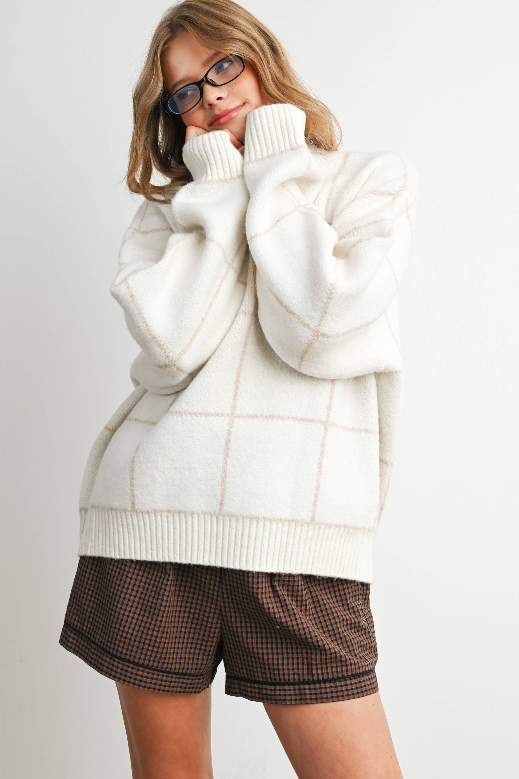 Woman wearing a white sweater over a checkered shirt with brown shorts on a light background