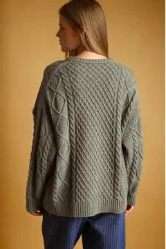 A woman shown from the back wearing an oversized cable-knit sweater in a muted sage green color, paired with dark blue pinstriped pants, set against a solid warm tan background.