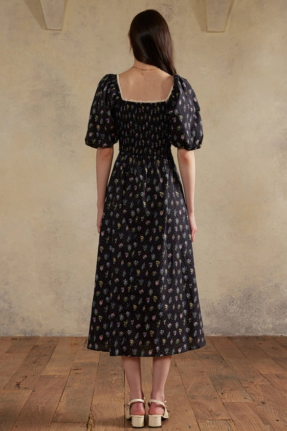 Woman wearing a black patterned dress standing against a beige wall.