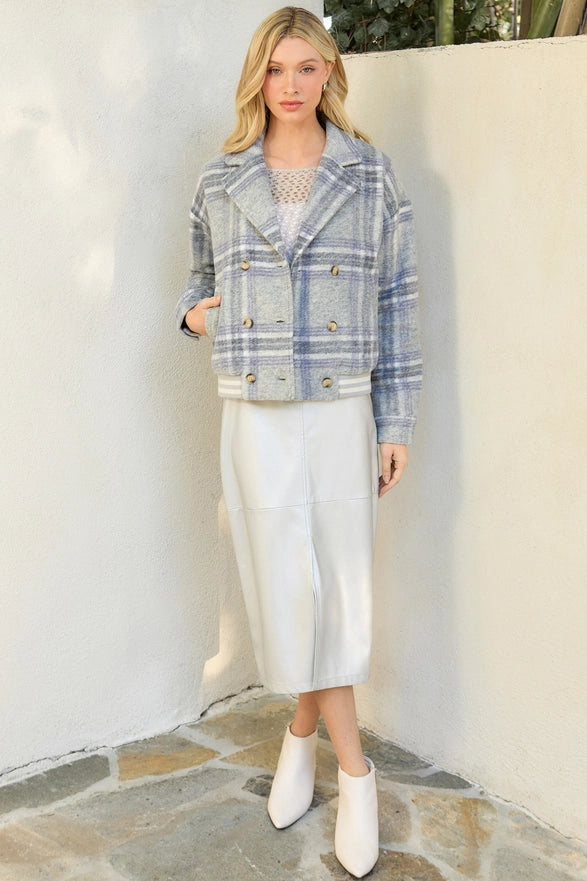 Woman wearing a plaid jacket, white skirt, and white boots against a white wall.