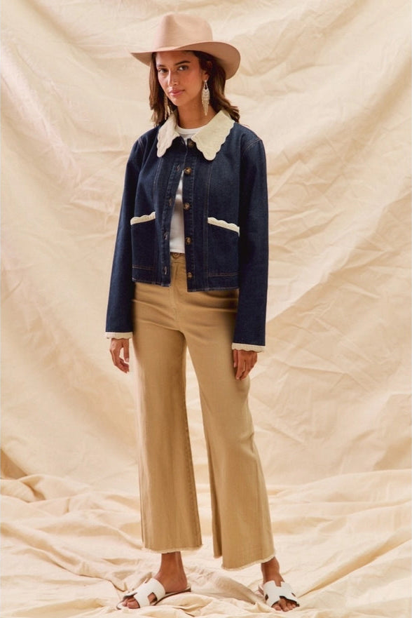 Woman wearing a denim jacket, beige pants, and a hat against a beige fabric background