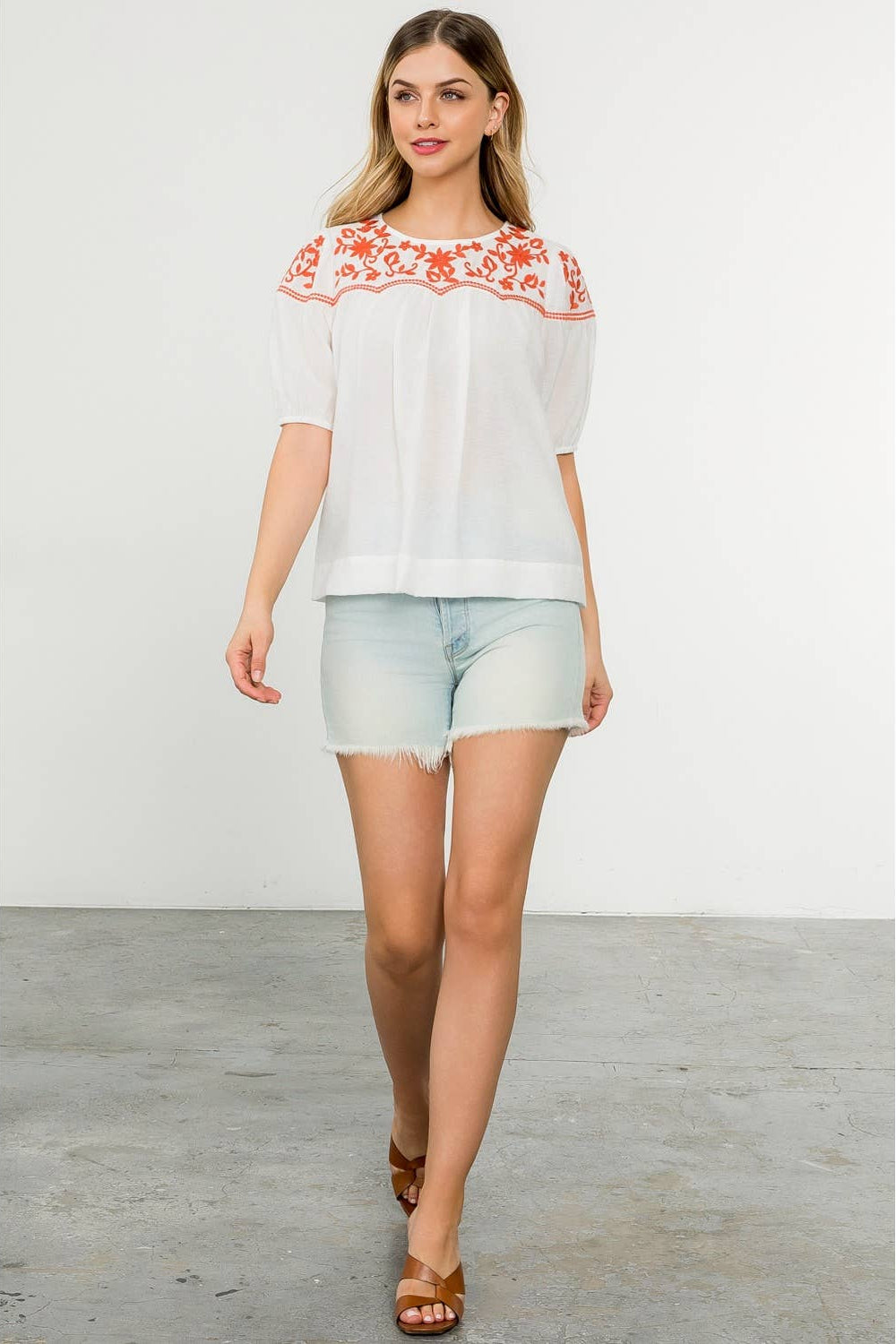 Woman wearing a white blouse with red floral embroidery and light blue shorts on a plain background
