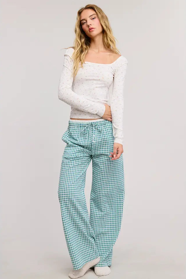 Woman wearing a white long-sleeve top and teal checkered pants on a plain background